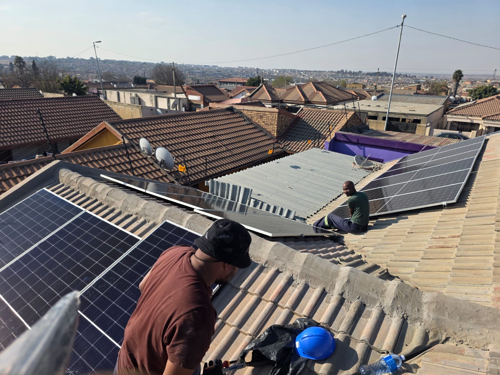 Two Kwevhos installers mounting solar panels on a tiled township roof, with rooftops stretching to the horizon behind them